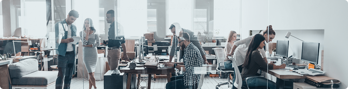 An open-plan office of people working at desks and monitors, seen through a glass partition.