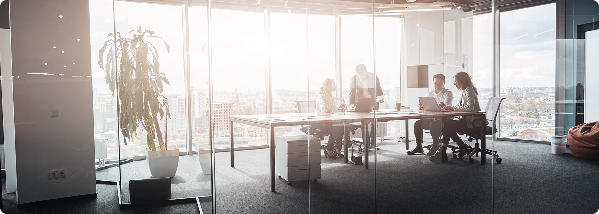 A meeting in a glass-walled conference room, four people around a table with a laptop, city skyline behind them.